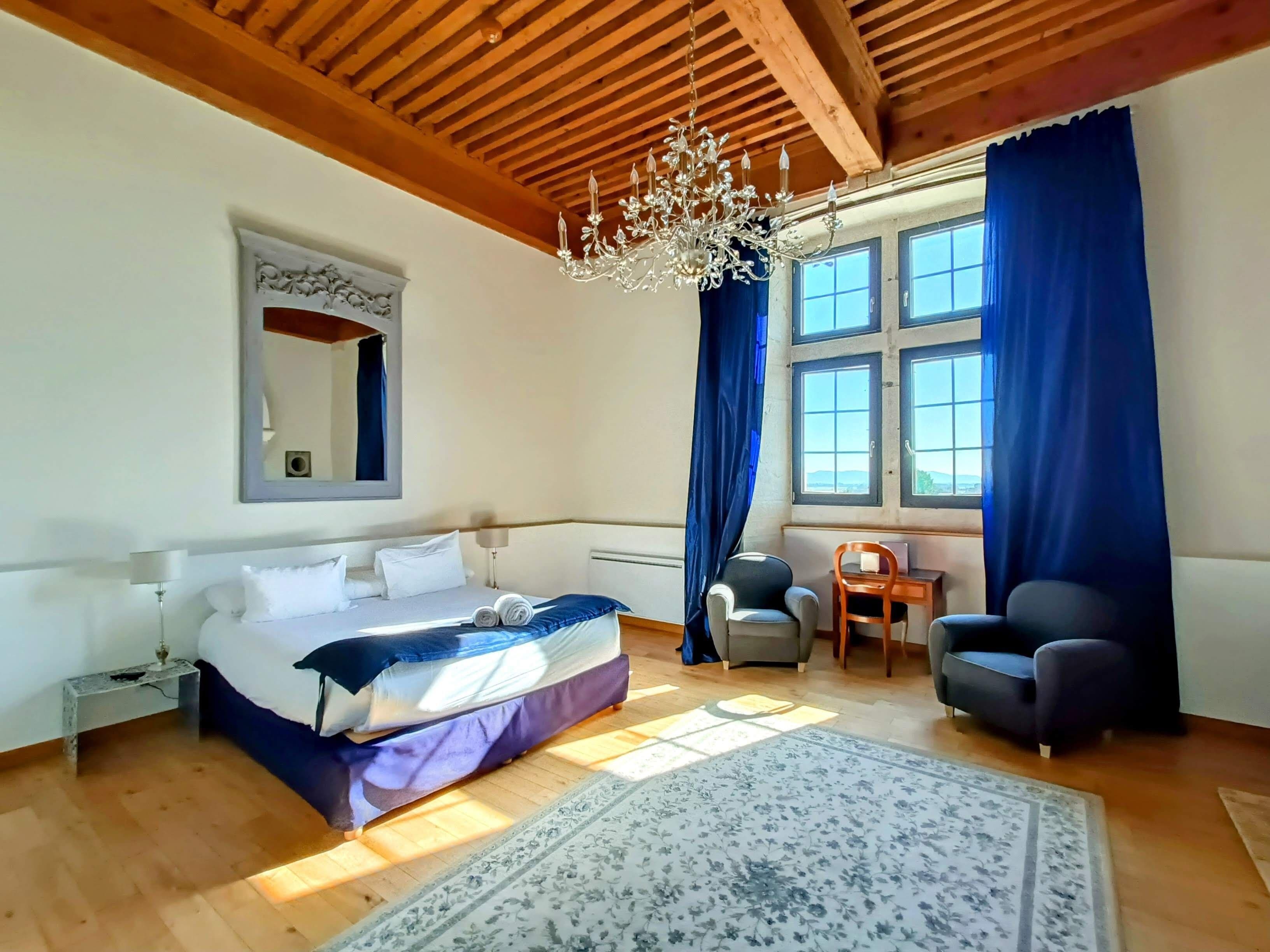 A bedroom with a bed, a desk, two chairs, a chandelier, and large windows with curtains.