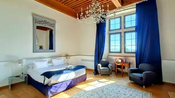 A bedroom with a bed, a desk, two chairs, a chandelier, and large windows with curtains.