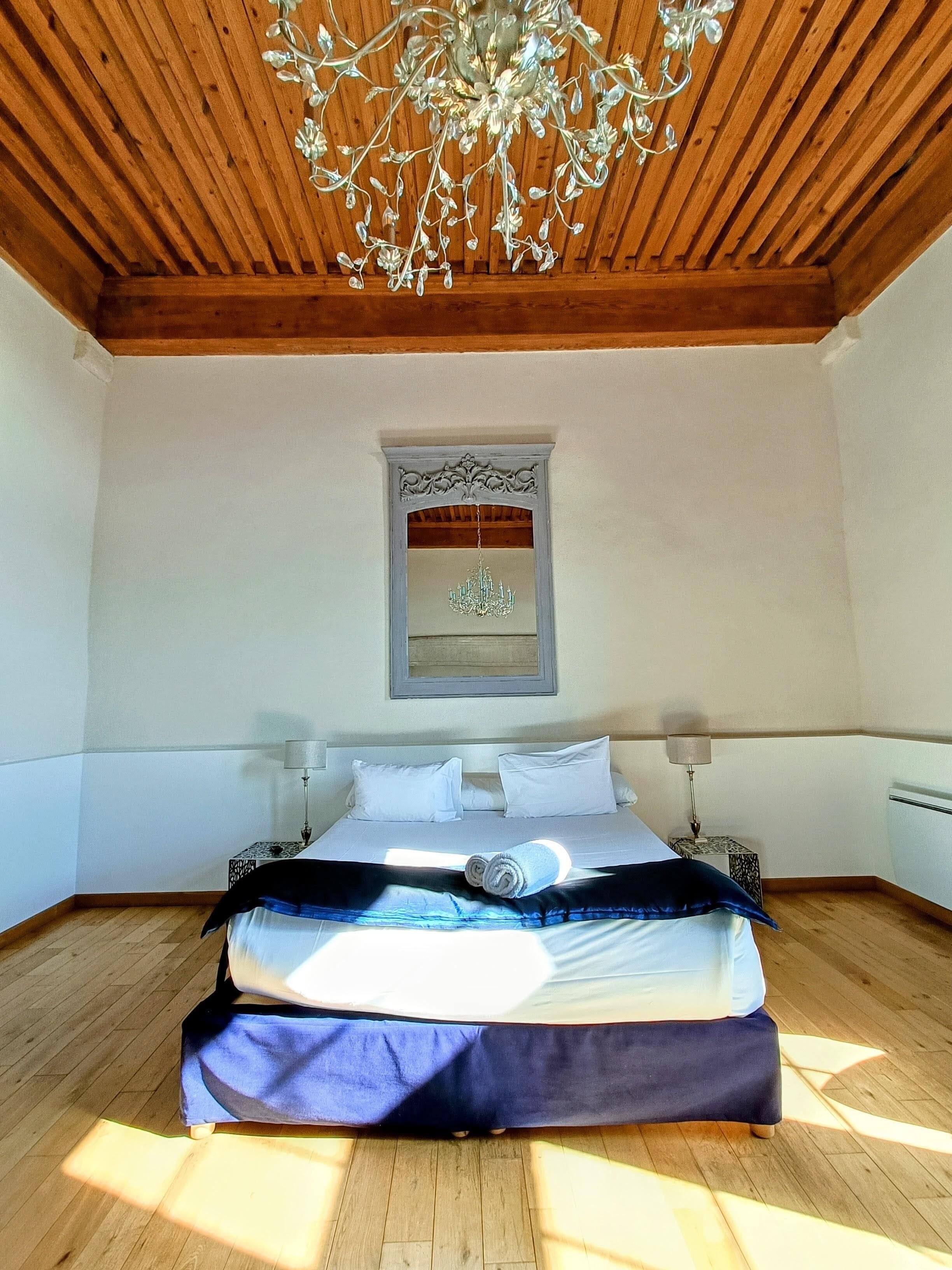A spacious bedroom with a wooden ceiling, a large bed with white and blue bedding, a chandelier, and a framed picture on the wall.