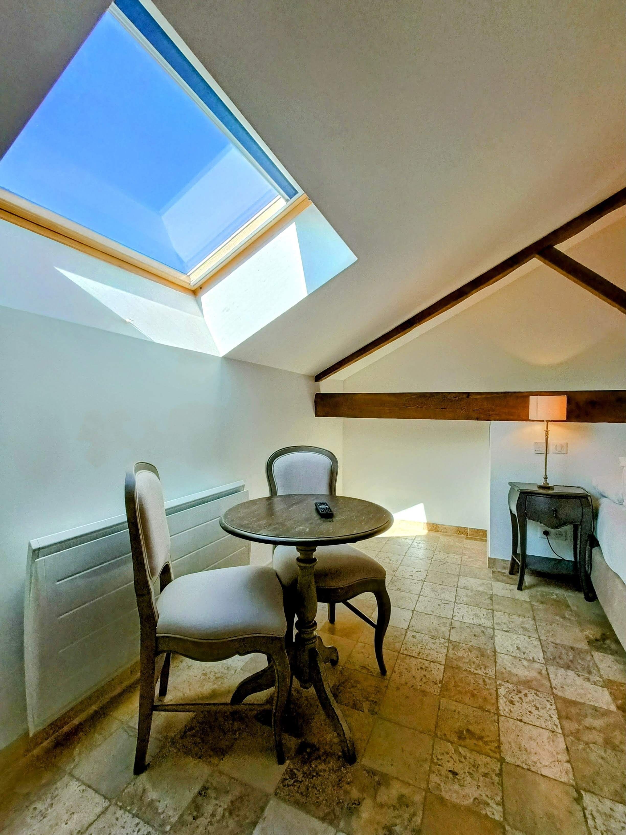 A room with a skylight, a round table with chairs, a lamp, and a bed.