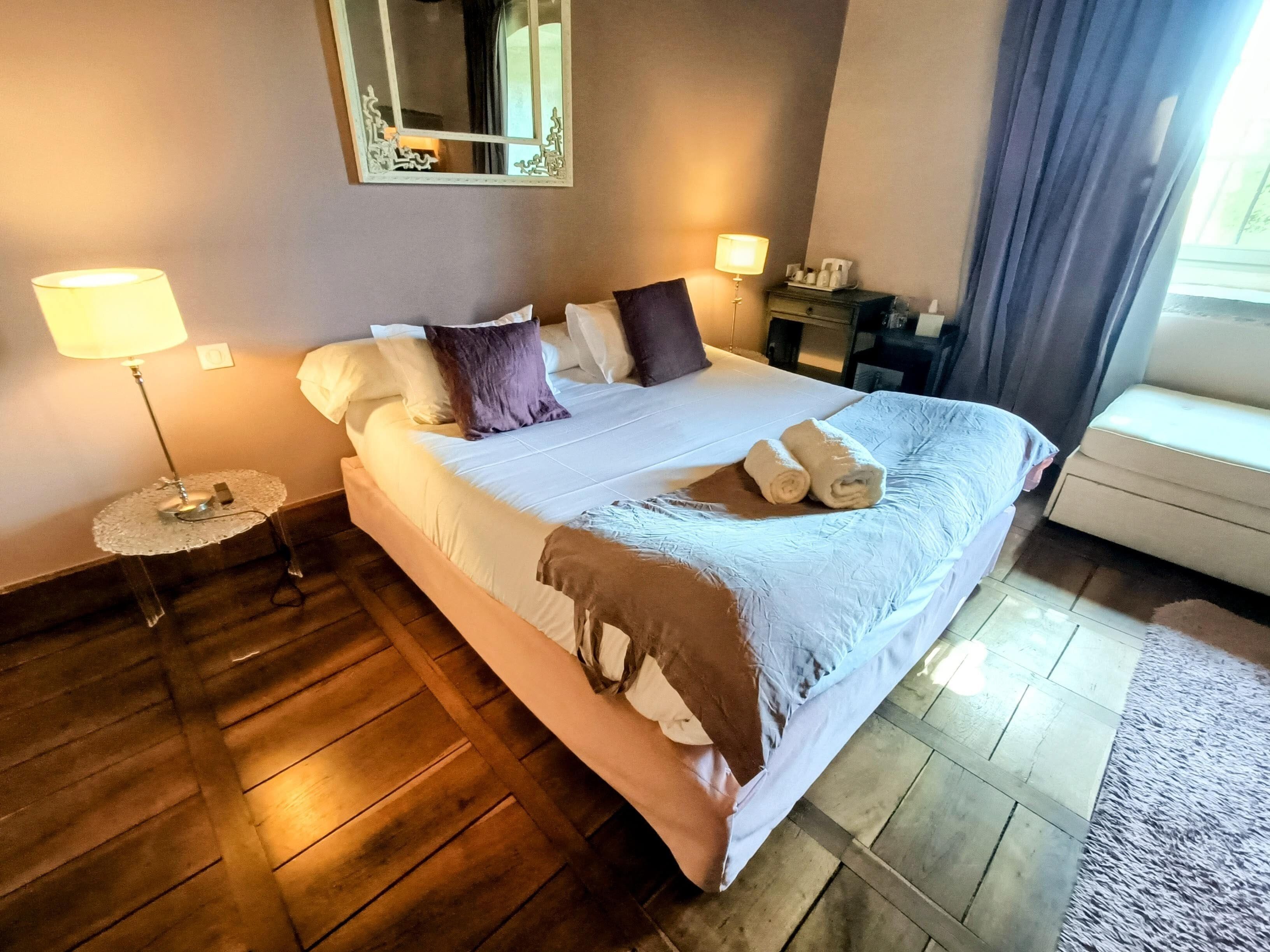 Una cama bien hecha con almohadas y una manta, una mesita de noche con lámpara, un espejo en la pared y una ventana con cortinas.