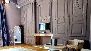 Een kamer met een houten visgraatvloer, een ronde tafel met een wit tafelkleed, twee witte fauteuils, een televisie op een schoorsteenmantel en een decoratieve spiegel.
