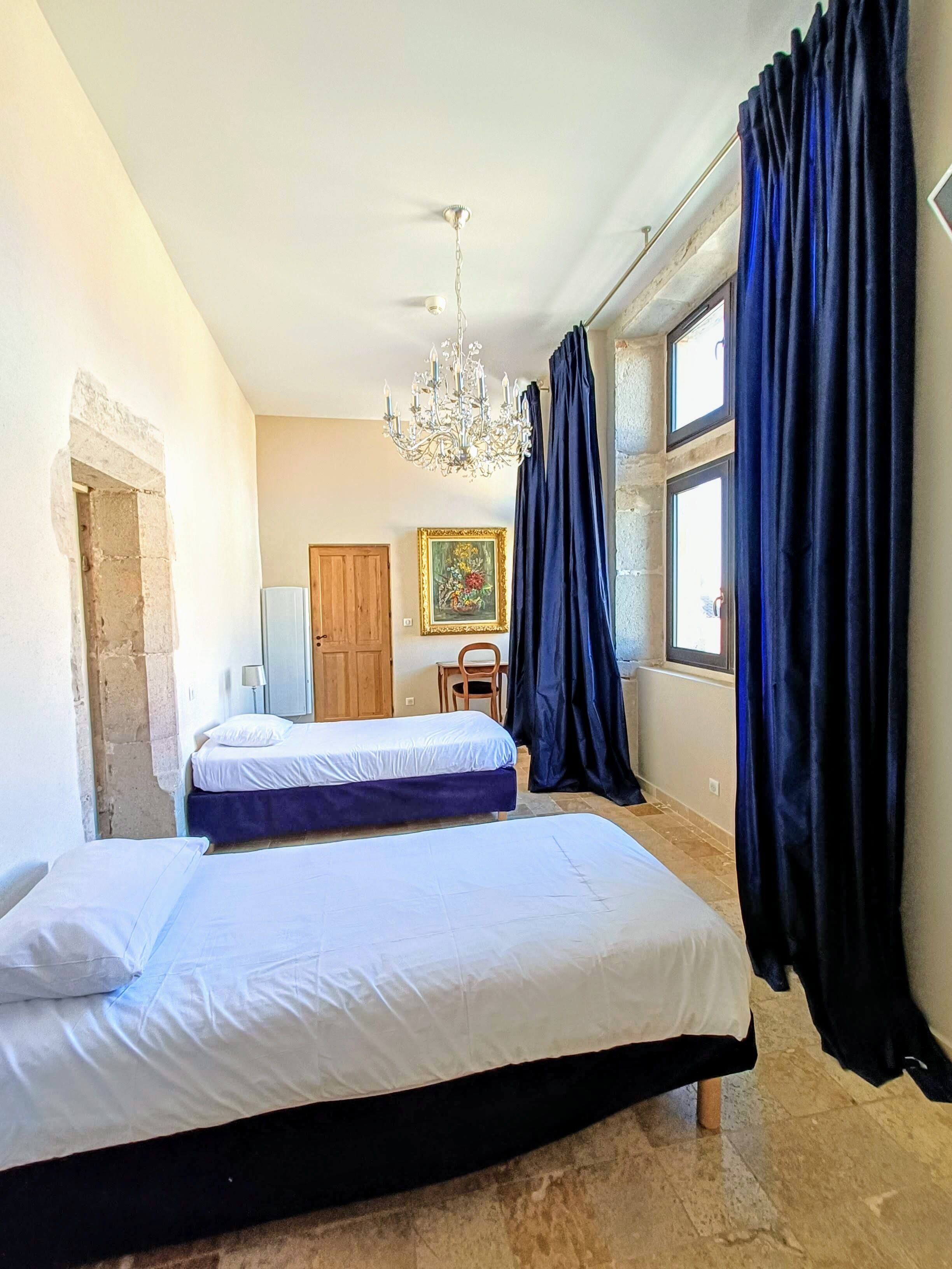 Une chambre avec deux lits, un lustre, une porte en bois, un tableau et de grandes fenêtres avec des rideaux.