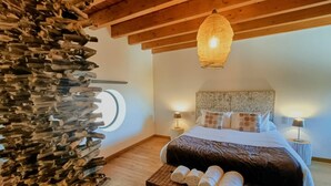Een slaapkamer met een houten plafond, een ronde spiegel, een bed met opgerolde handdoeken en twee bedlampjes.