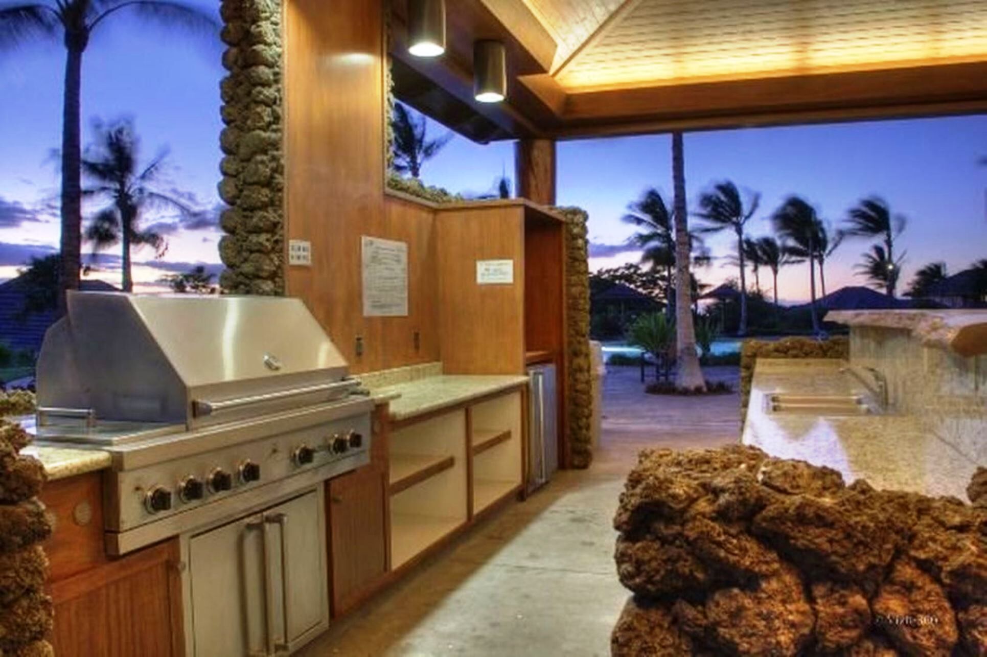 Une cuisine extérieure équipée d’un barbecue, de meubles en bois et offrant une vue sur des palmiers et un coucher de soleil.