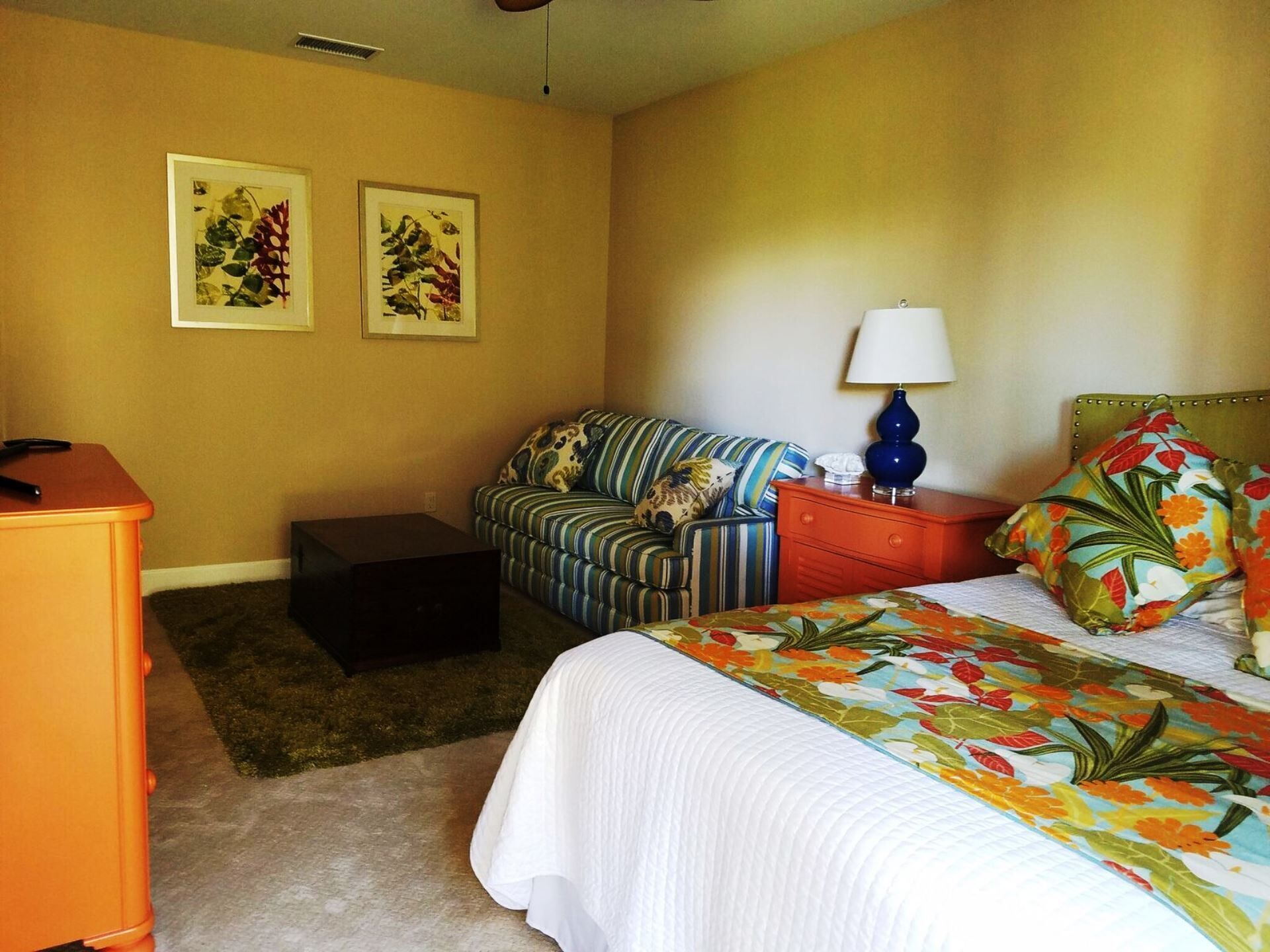 A hotel room with a bed, a striped sofa, a small table, a nightstand, a lamp, and two framed botanical prints on the wall.
