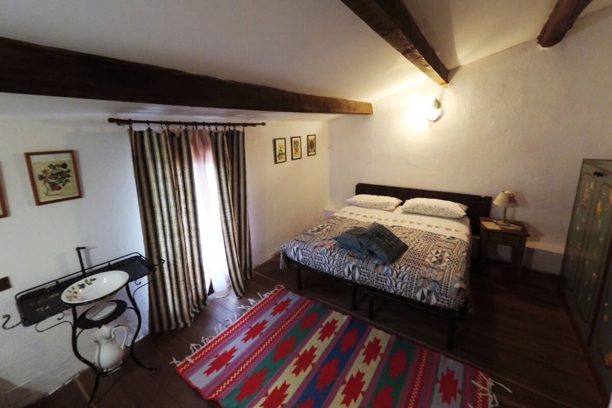 A bedroom with a bed, a nightstand, a bedside table, a window with curtains, a radiator, and a wall-mounted light.