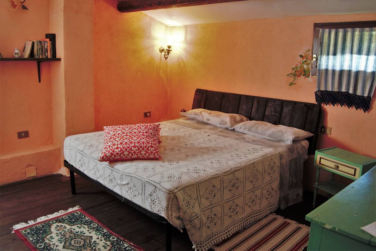 A bedroom with a bed, a nightstand, a wall-mounted lamp, a shelf with books, and a window with curtains.