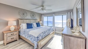 Une chambre à coucher comprenant un lit, une table de chevet, une commode, un ventilateur de plafond et de grandes fenêtres offrant une vue dégagée.