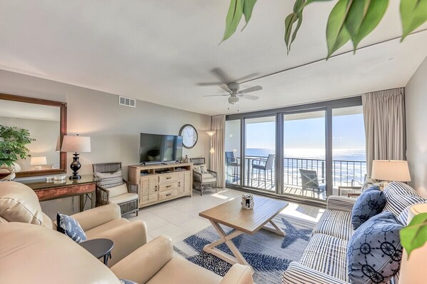 A spacious living room with a large window, a ceiling fan, and a balcony with a view.