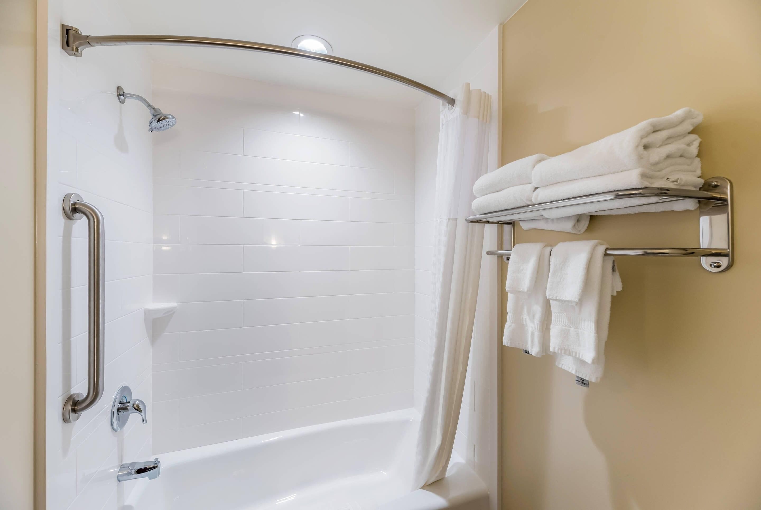 A modern bathroom with a white bathtub, a shower with a handheld showerhead, a grab bar, and a towel rack with folded towels.