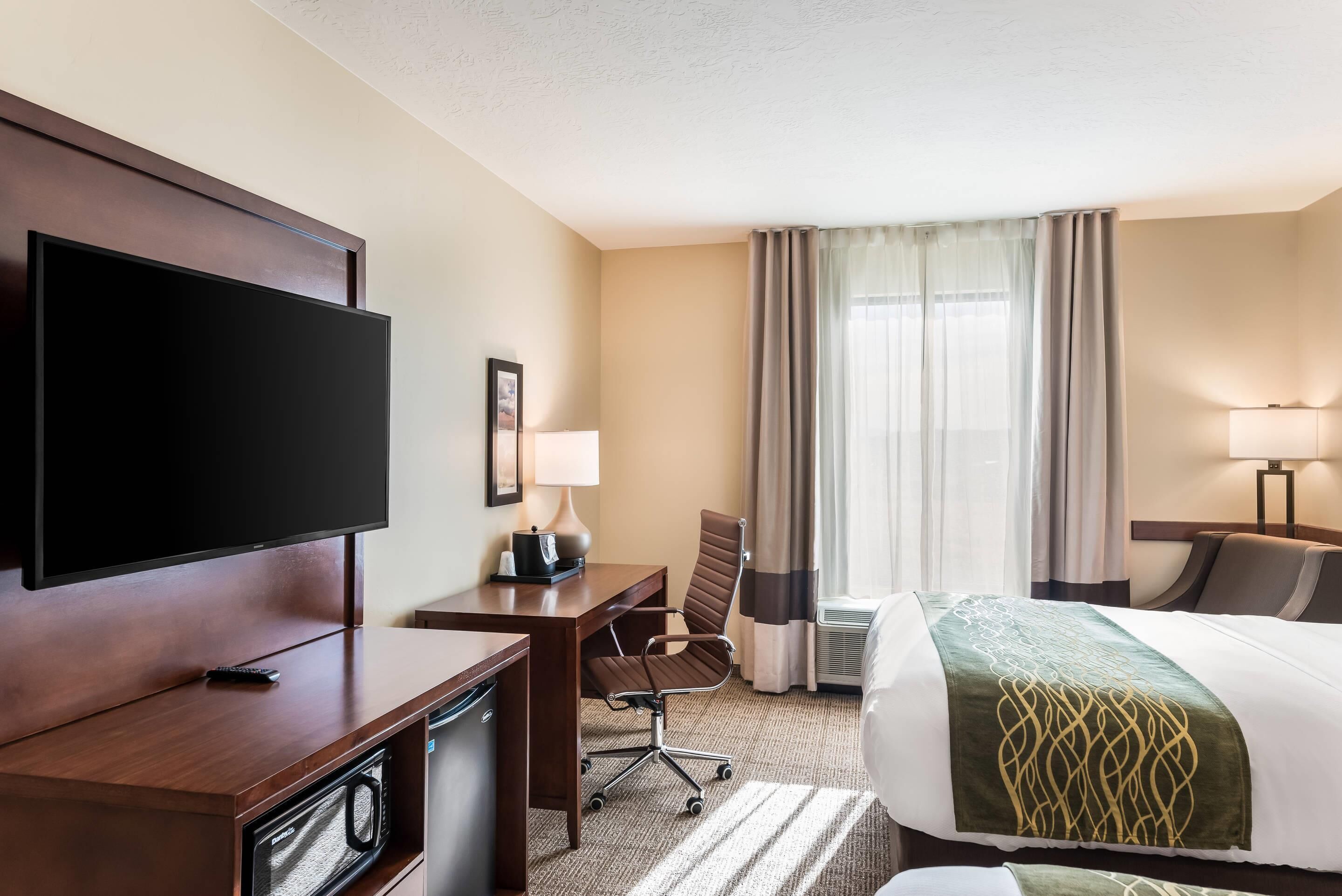 A hotel room with a flat-screen TV, a desk with a lamp, a chair, a bed with a green blanket, and a window with curtains.