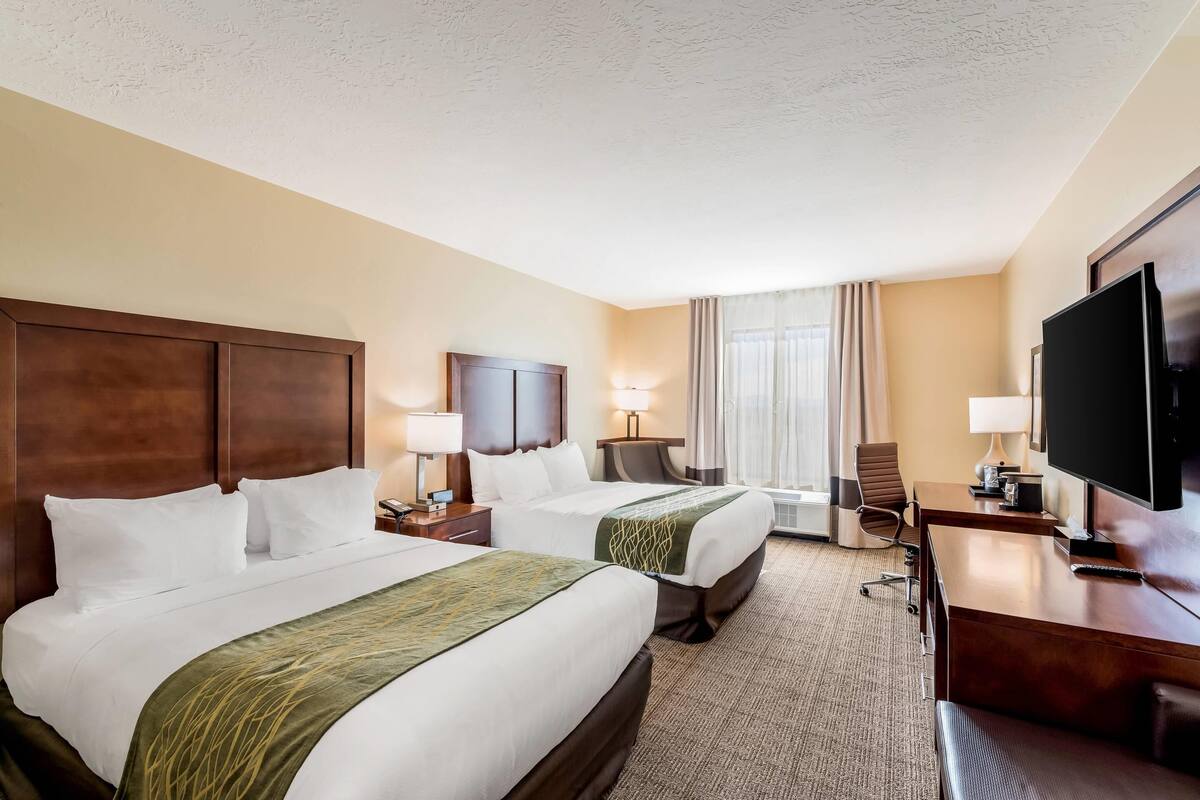 A hotel room with two beds, a desk, a TV, and a window with curtains.