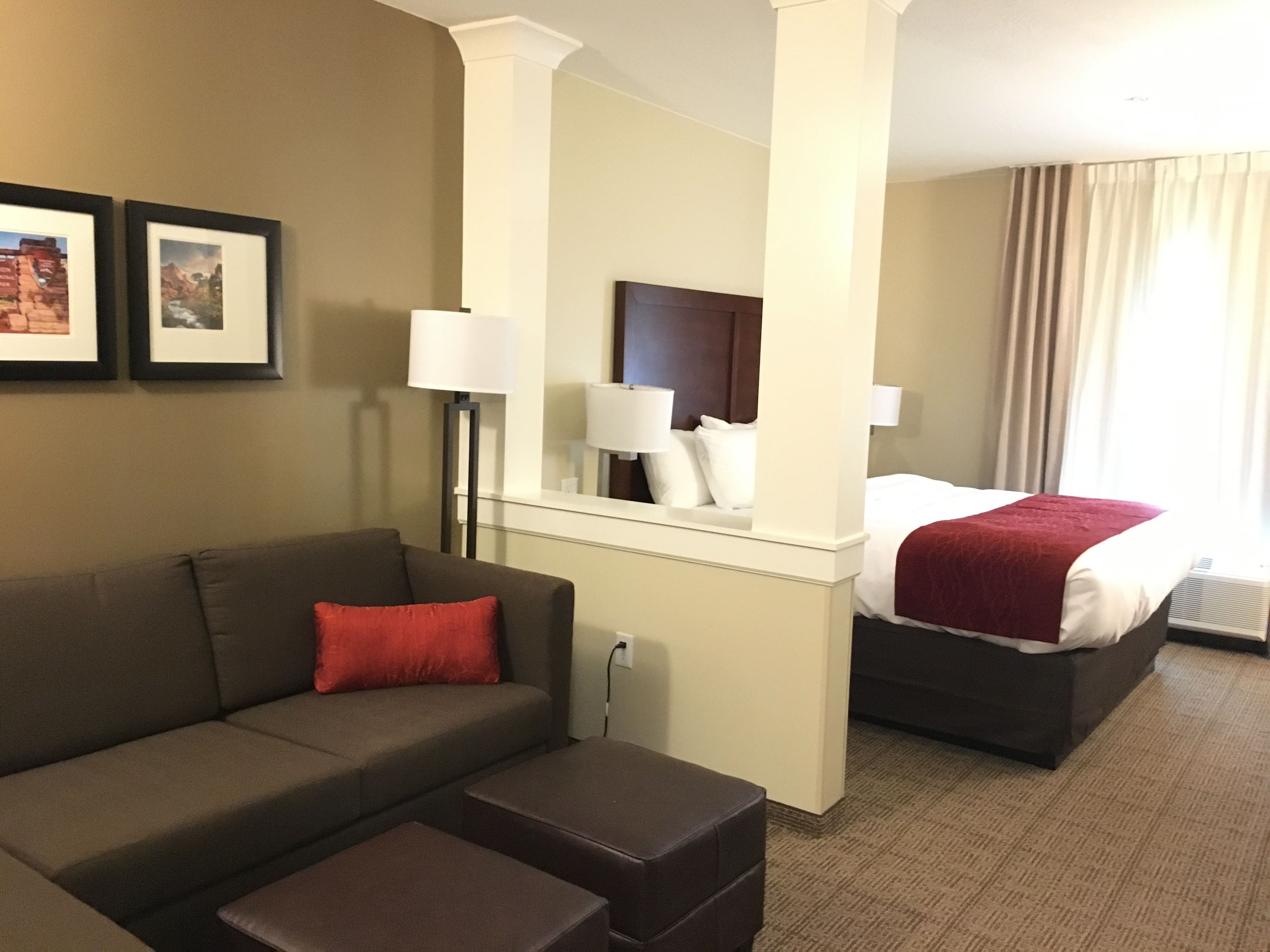 Una habitación de hotel con sofá, un pequeño reposapiés, cama con manta roja, mesita de noche y espejo.