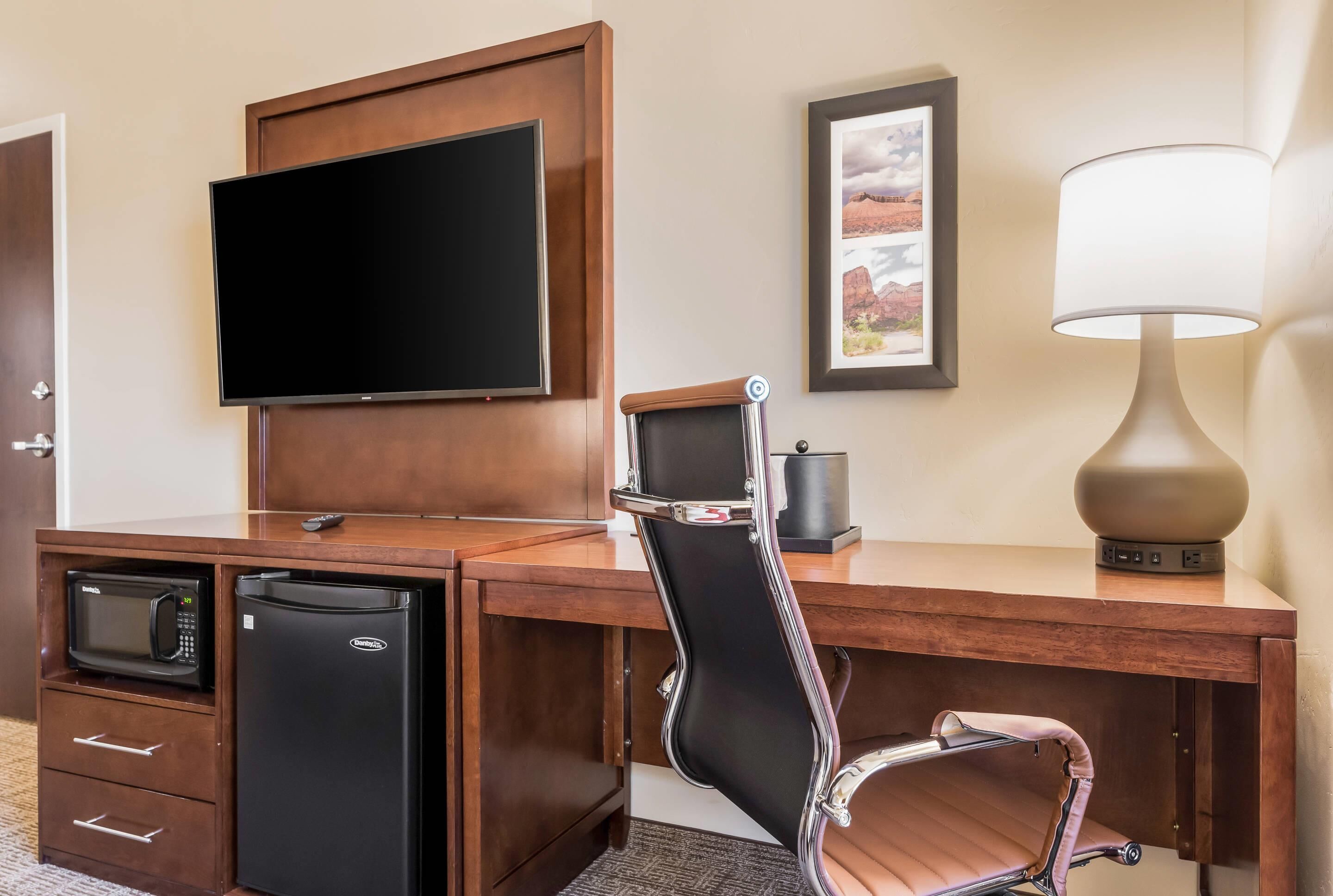 A hotel room with a wooden desk, a flat-screen TV, a refrigerator, a microwave, a chair, a lamp, and a framed picture on the wall.