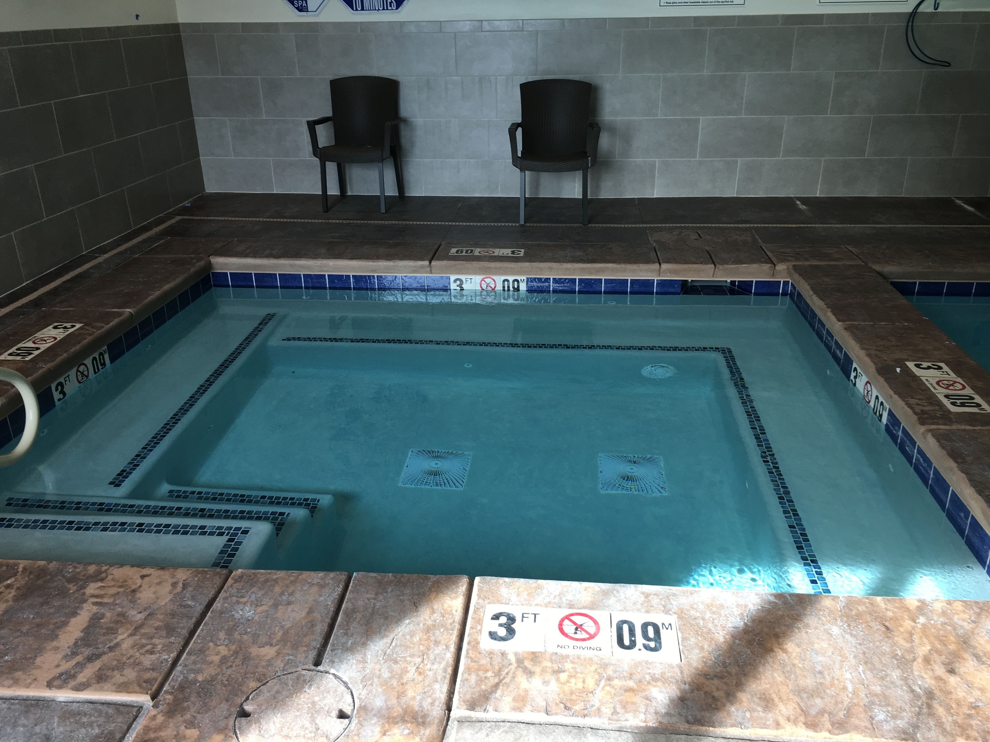 Piscina con piastrelle blu, due sedie nere e un muro rivestito di piastrelle con un cartello.