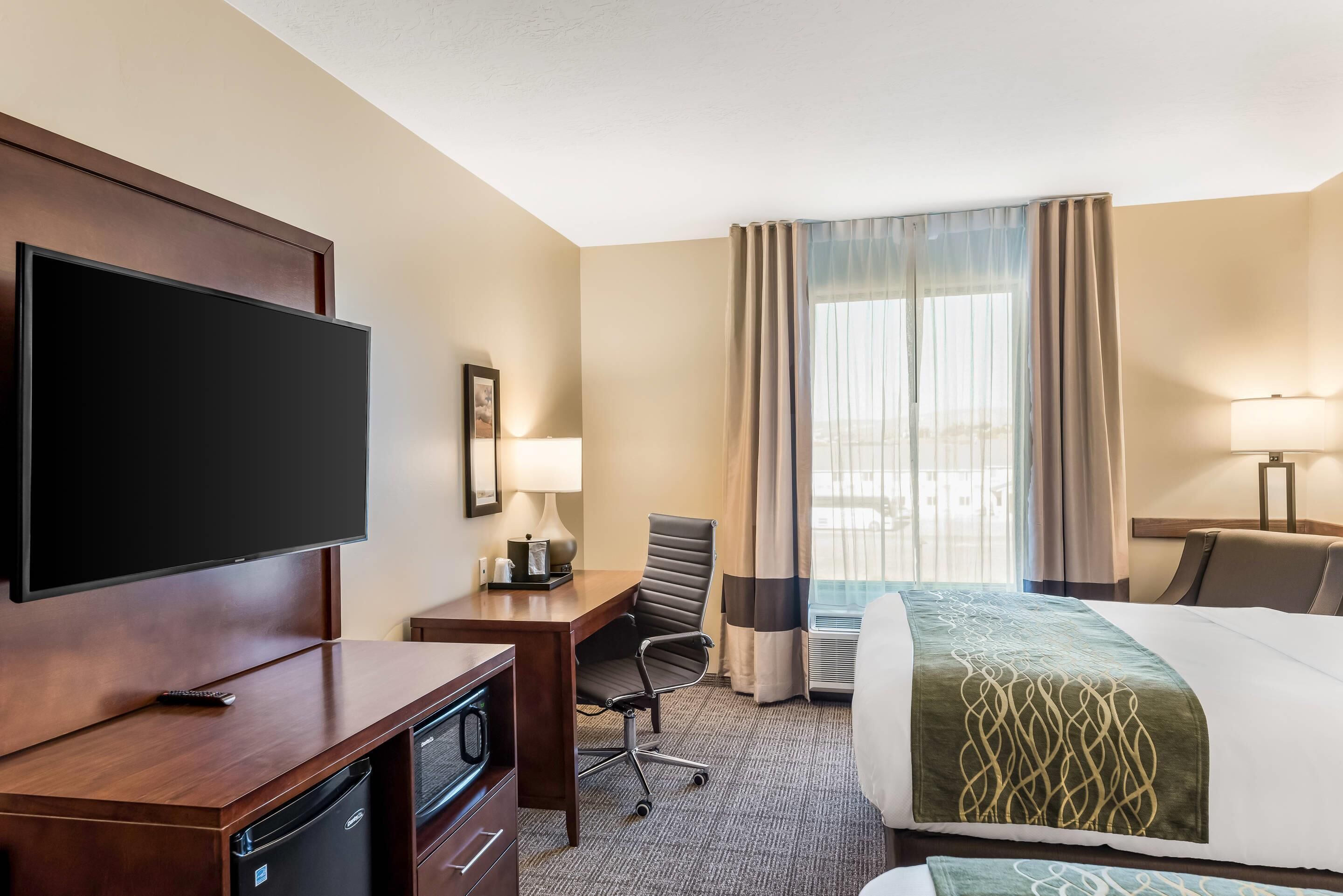 Ein Hotelzimmer mit Flachbildfernseher, Schreibtisch mit Stuhl, Bett mit grüner Decke und einem Vorhangfenster.