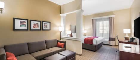 A hotel room with a sofa, ottoman, a bed, a desk, a TV, and framed pictures on the wall.