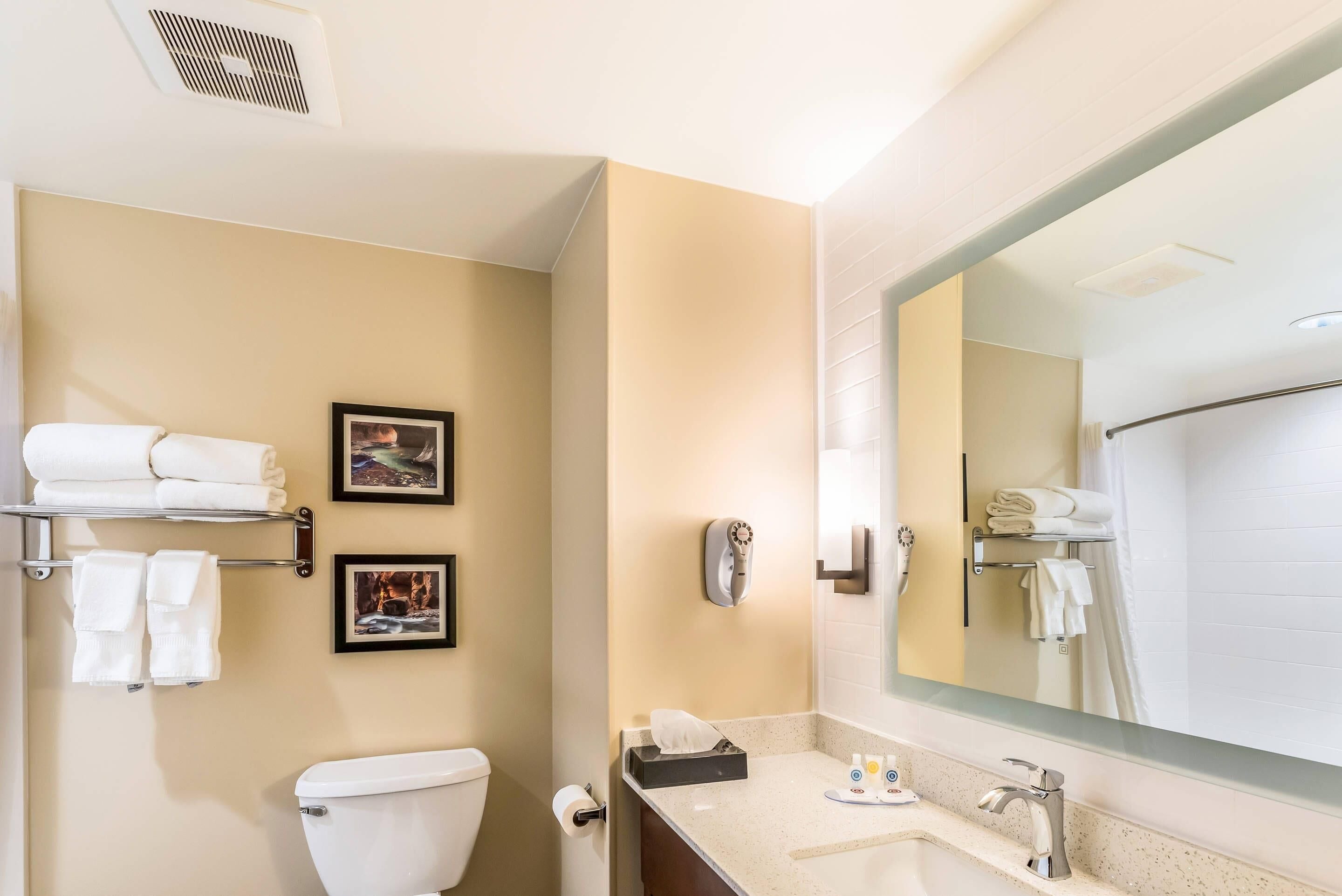Un baño moderno con inodoro, lavamanos y un espejo grande. Hay toallas en un perchero y un cuadro en la pared.