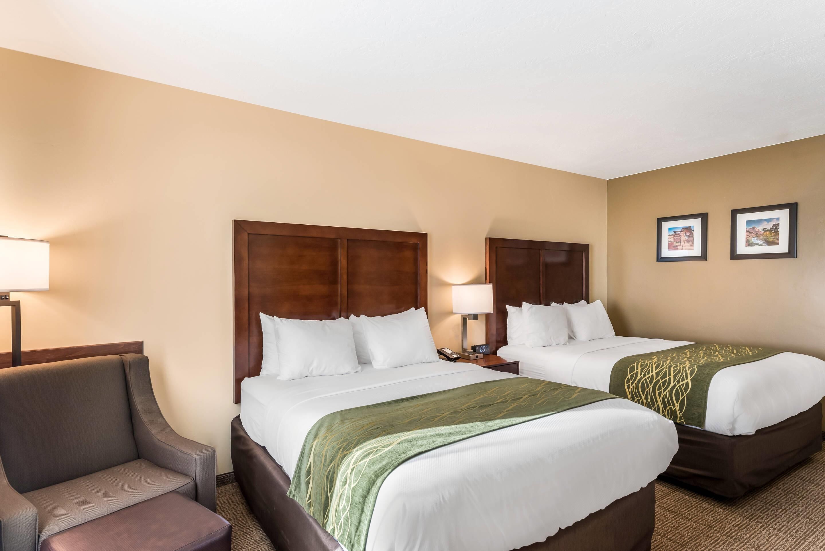 A hotel room with two beds, a chair, a nightstand with a lamp, and two framed pictures on the wall.