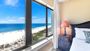 Ein Hotelzimmer mit großem Fenster und Blick auf den Strand, einem Nachttisch mit Lampe sowie einem Bett mit gemustertem Kissen.