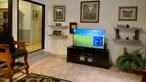 A living room with a large flat-screen TV on a black stand, a wooden chair, a patterned rug, and a potted plant. - Golfers getaway - putting green, billiards, bar, arcade , poker table and more (Pinehurst)