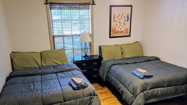 A bedroom with two beds, a nightstand, a lamp, and a window with blinds. - Golfers getaway - putting green, billiards, bar, arcade , poker table and more (Pinehurst)