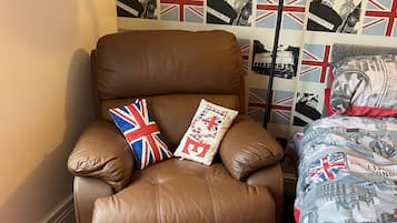 Un fauteuil en cuir marron agrémenté de deux coussins, dont l’un arbore un motif Union Jack, dans une pièce dotée d’un tapis rouge et d’un mur décoré d’un papier peint sur le thème britannique.