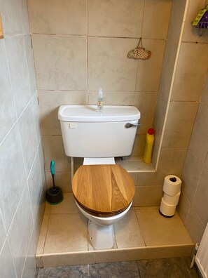 A small bathroom with a toilet, a wooden seat, a plunger, a toilet brush, and a roll of toilet paper.