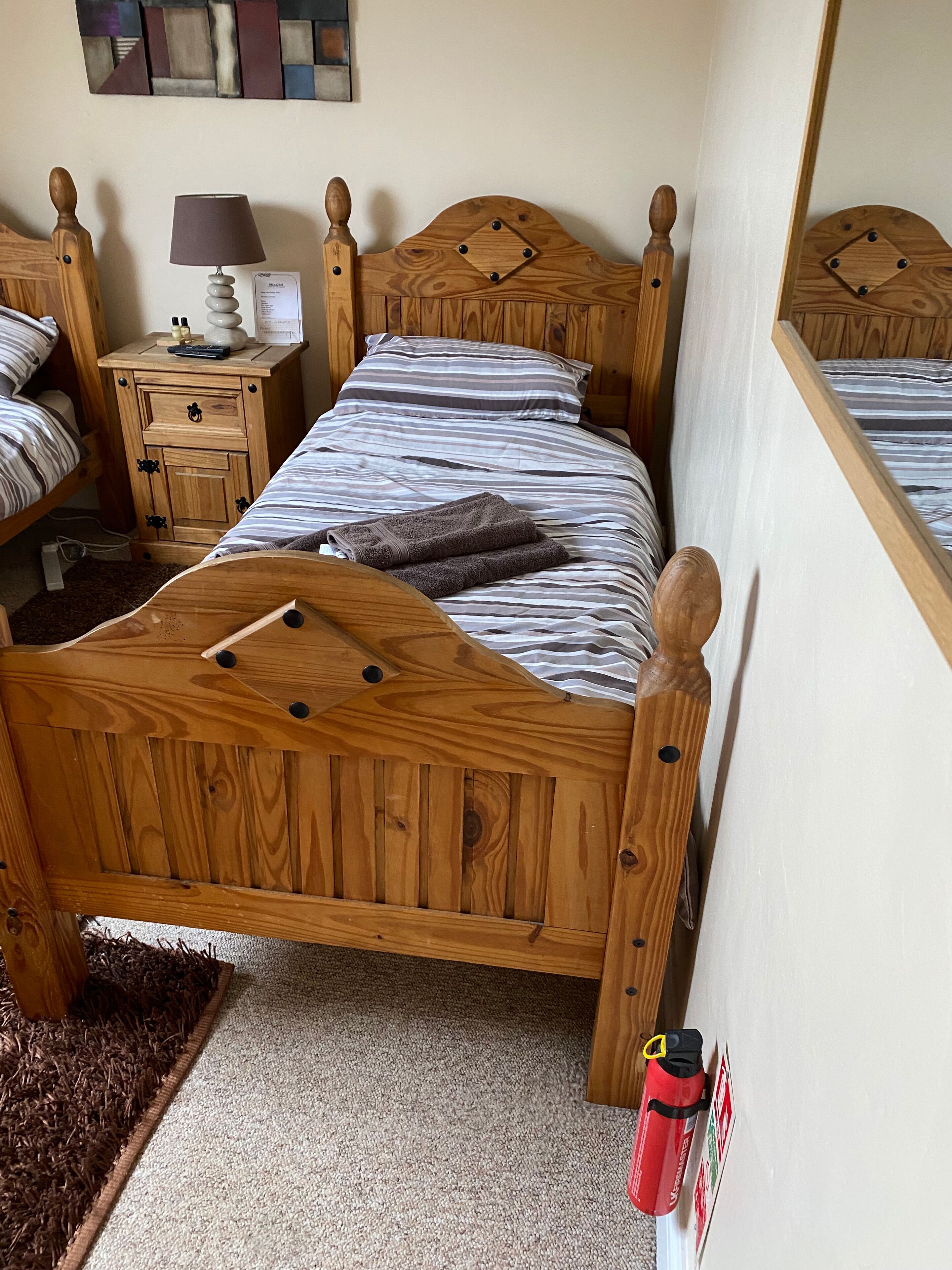 A wooden bed with a headboard, a bedside table with a lamp, and a red water bottle on the floor.