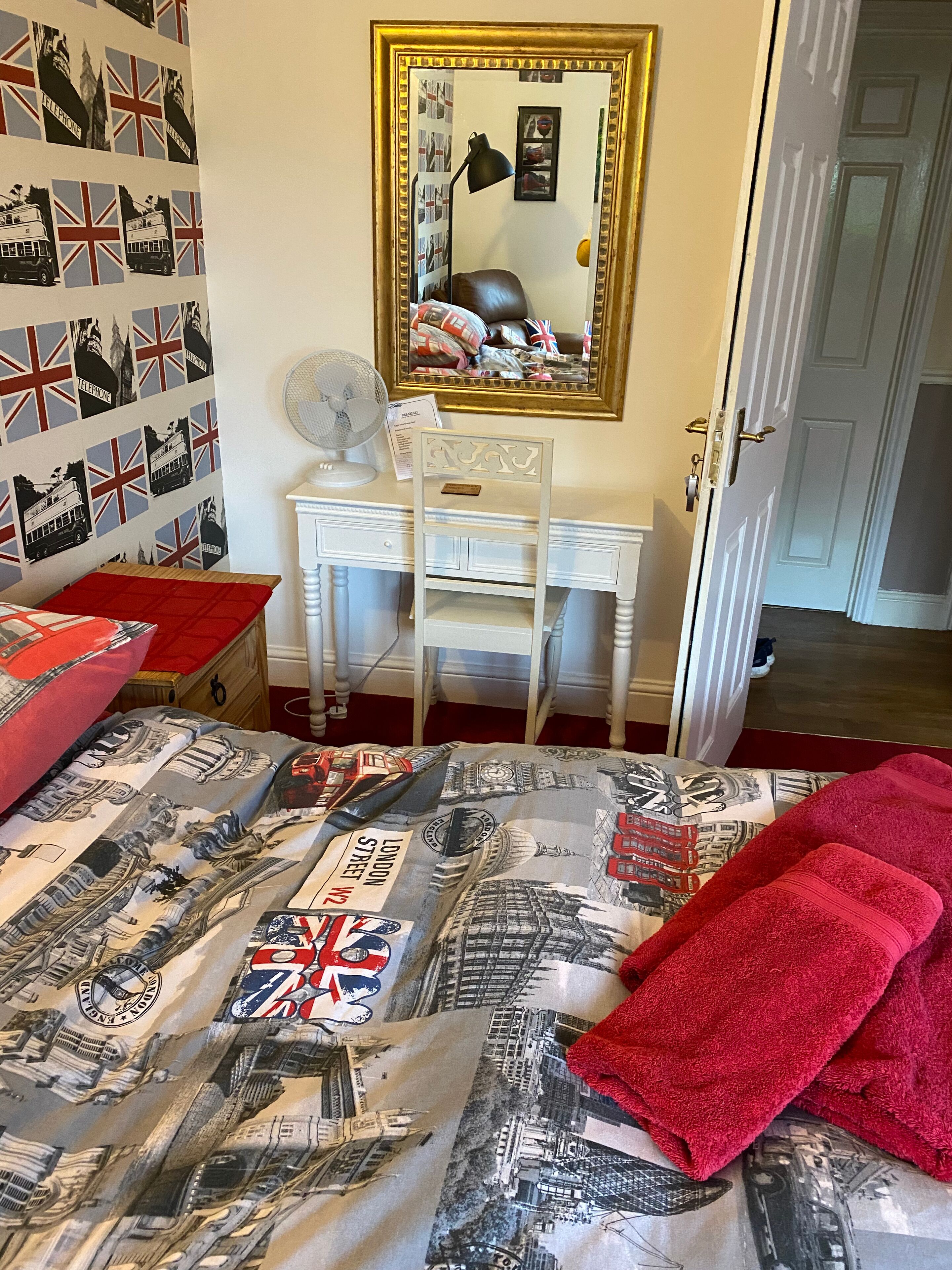 Ein Schlafzimmer mit einem Bett, einem Schreibtisch und einem Stuhl. An der Wand hängt ein Spiegel und zur Rechten befindet sich eine Tür.