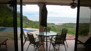 A glass-enclosed patio with a table and chairs, overlooking a pool and lush greenery. - Sublime Obsession-Amazing Views & Sunsets on the Pacific Ocean (Coyote)