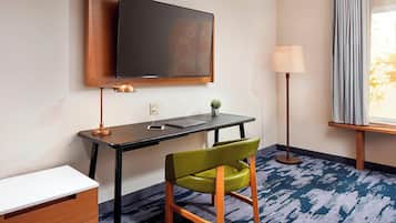 Une chambre d’hôtel équipée d’un téléviseur à écran plat fixé au mur, d’un bureau avec un ordinateur portable, d’un fauteuil vert, d’une lampe et d’une fenêtre avec des rideaux.