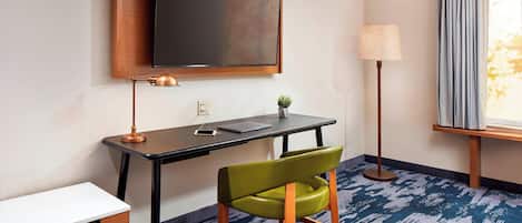 A hotel room with a flat-screen TV mounted on the wall, a desk with a laptop, a green chair, a lamp, and a window with curtains.