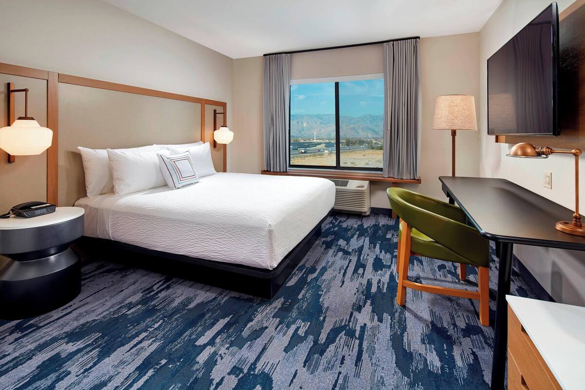 A hotel room with a bed, a desk, a chair, a TV, and a window with a mountain view.