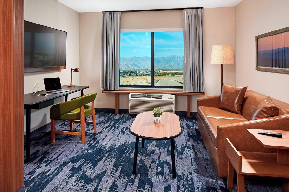 A hotel room with a large window, a desk with a laptop, a green chair, a brown leather sofa, and a small table with a plant.