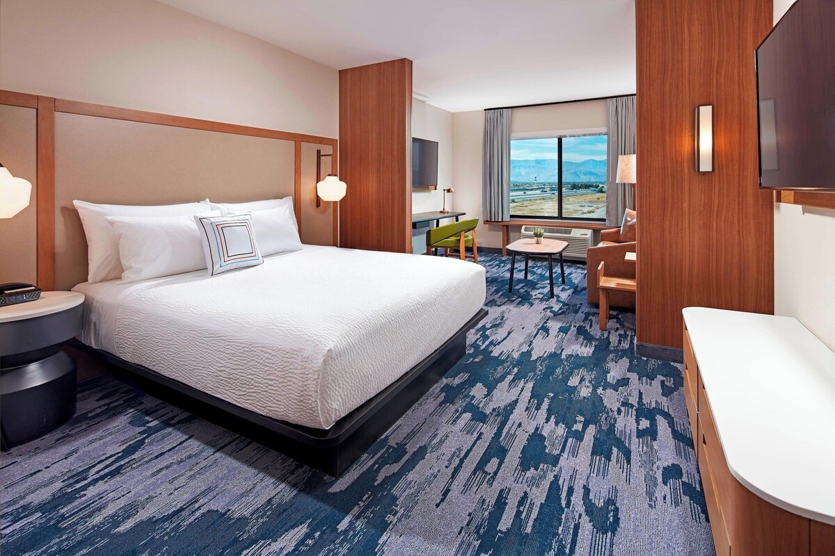 A hotel room with a large bed, a desk, a chair, a TV, and a window with a view.