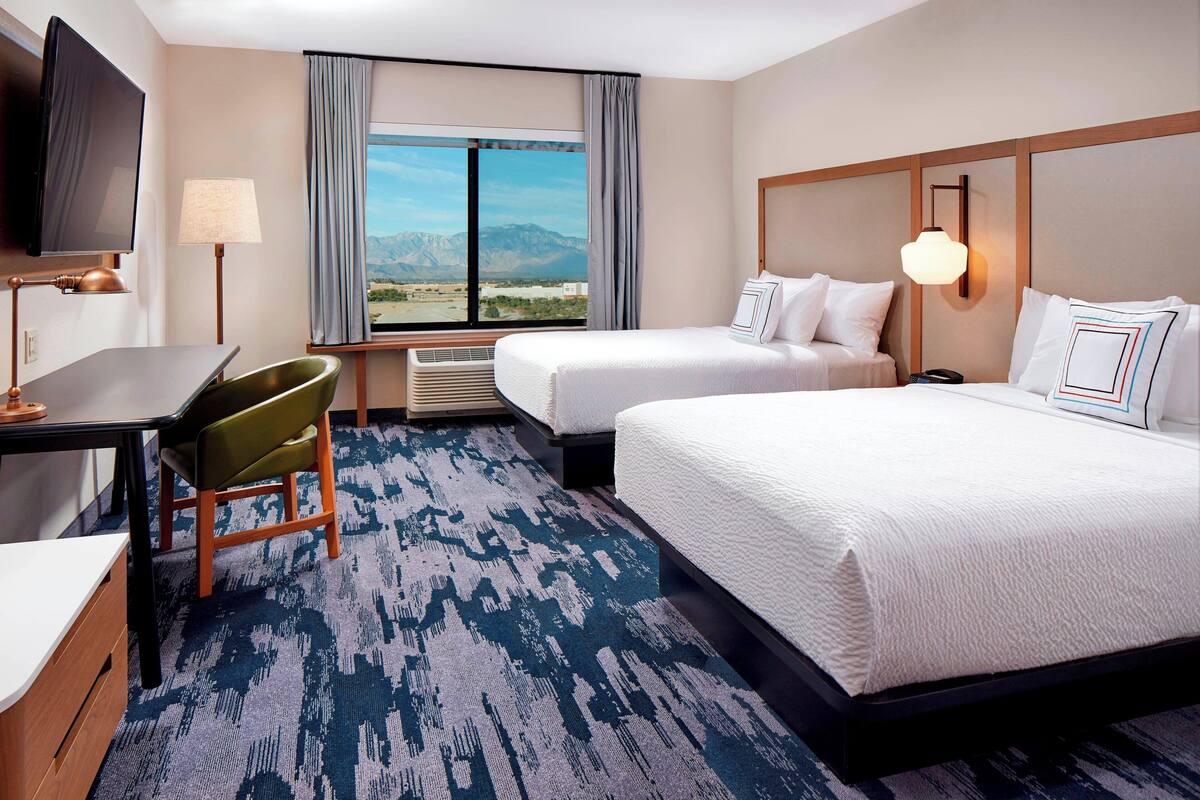 A hotel room with two beds, a desk, a chair, a TV, and a large window with a mountain view.