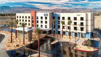 A multi-story hotel with a modern design, featuring a parking lot with palm trees and a view of mountains in the background.