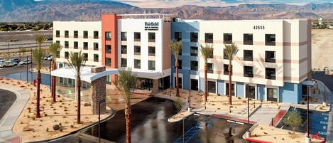 A multi-story hotel with a modern design, featuring a parking lot with palm trees and a view of mountains in the background.