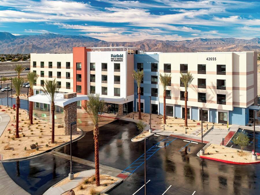 A multi-story hotel with a modern design, featuring a parking lot with palm trees and a view of mountains in the background.