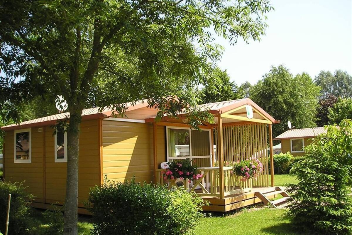 Una casa rodante amarilla con porche, rodeada de vegetación y una pequeña terraza.