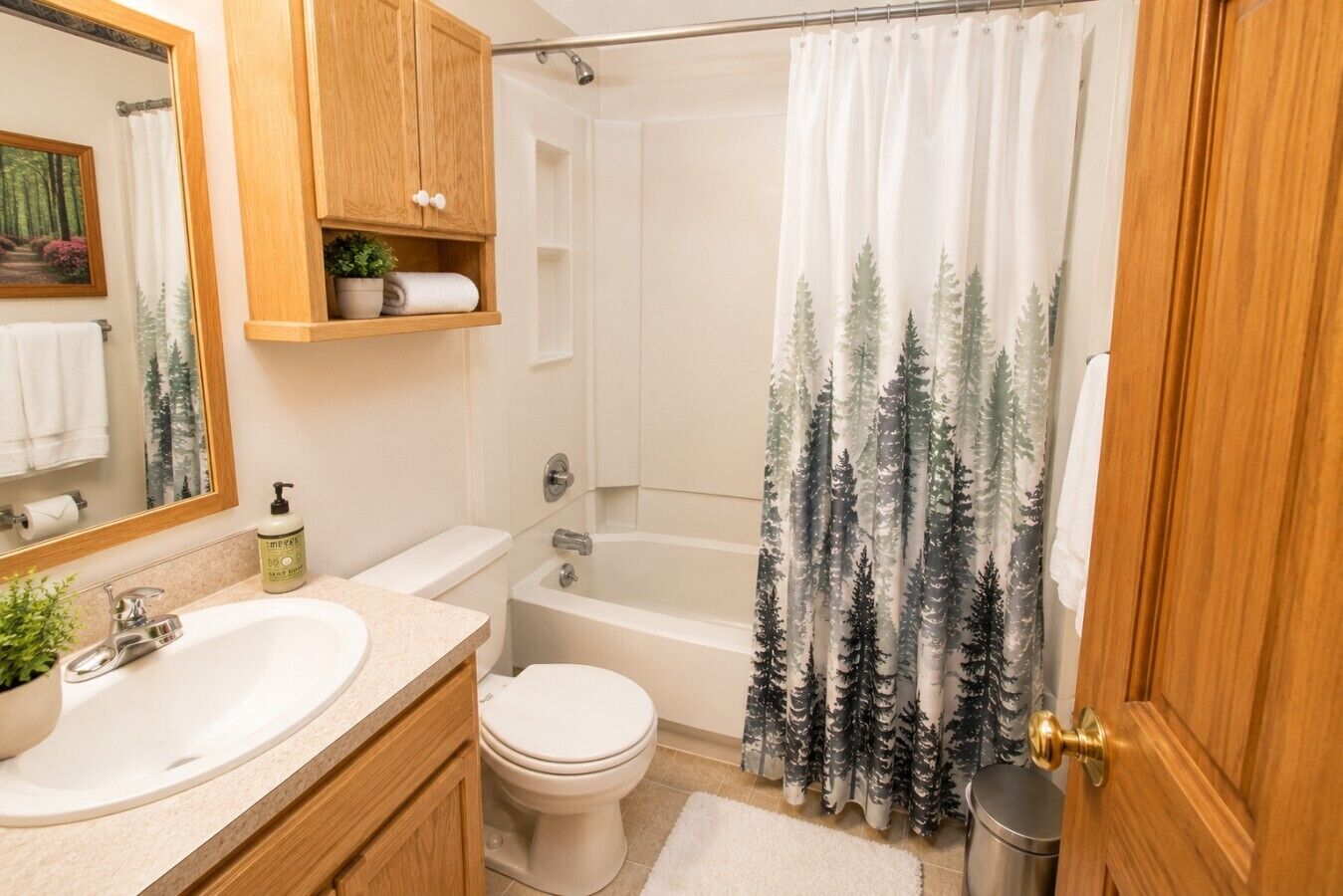 Un baño con un mueble de madera, un lavabo, un inodoro, una bañera y una cortina de ducha con estampado de bosque.