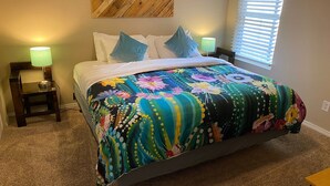 A bedroom with a bed, two nightstands, a lamp, a wooden headboard, a floral bedspread, and a chevron-patterned wall art piece. - ABQ Uptown Condo near I-40, 2bd/1ba, AC! (Albuquerque)