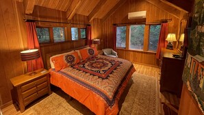 A cozy bedroom with a wooden ceiling, a large bed with patterned bedding, a nightstand, a chair, a dresser, and a window with curtains. - Gramma's 4BR Home, Lake, Wooded, Hot Tub, Swim, Swings, Gated Estate, Gorgeous (Friday Harbor)