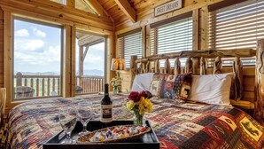 Ein Zimmer in einer Holzhütte mit einem großen Bett, einem Tablett mit Weingläsern und Pralinen, einer Vase mit Blumen und einem Schild mit der Aufschrift 'SWEET DREAMS'.