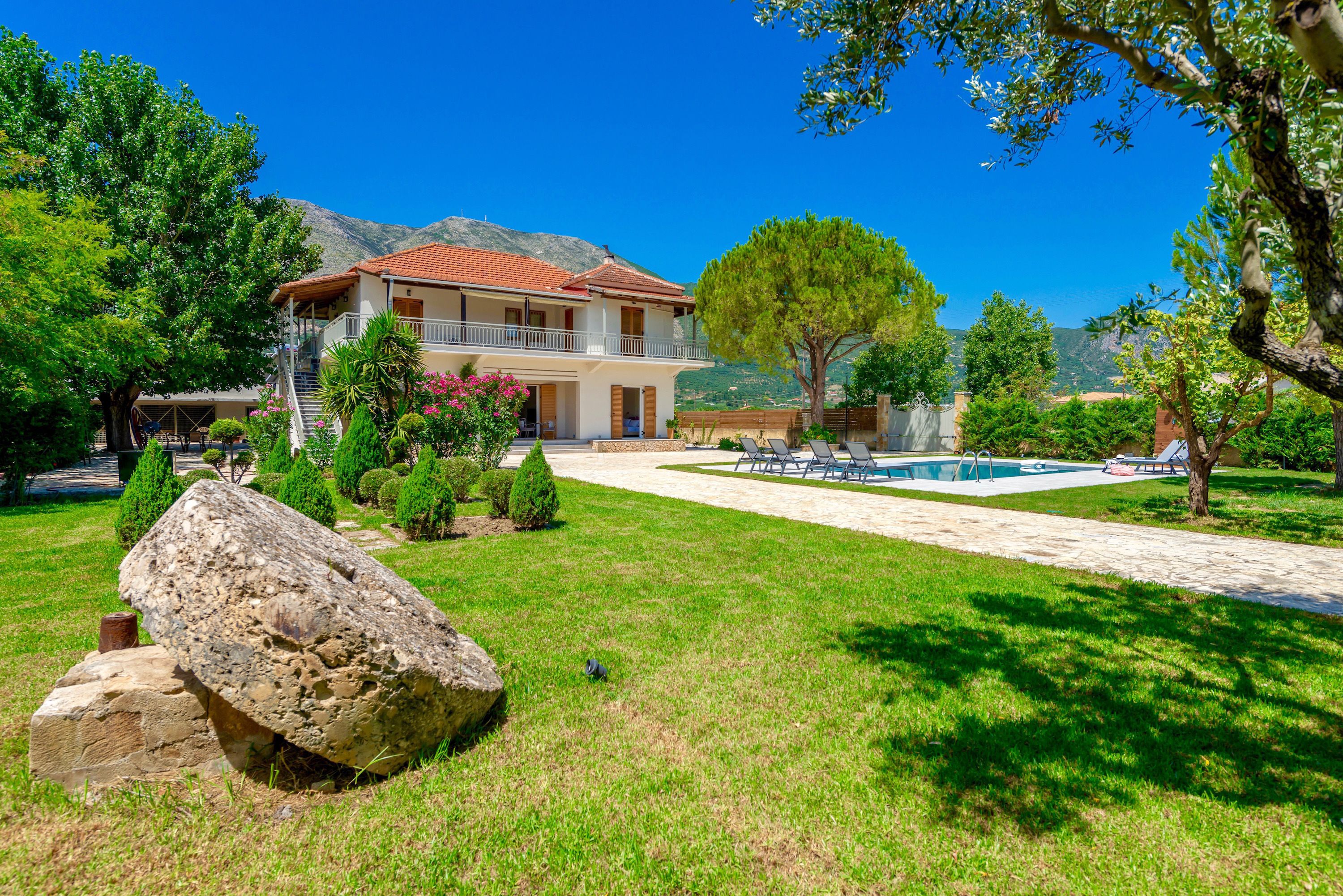 A two-story house with a balcony, a swimming pool, and a mountainous backdrop.