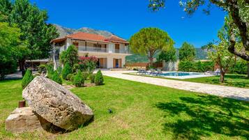 A two-story house with a balcony, a swimming pool, and a mountainous backdrop.