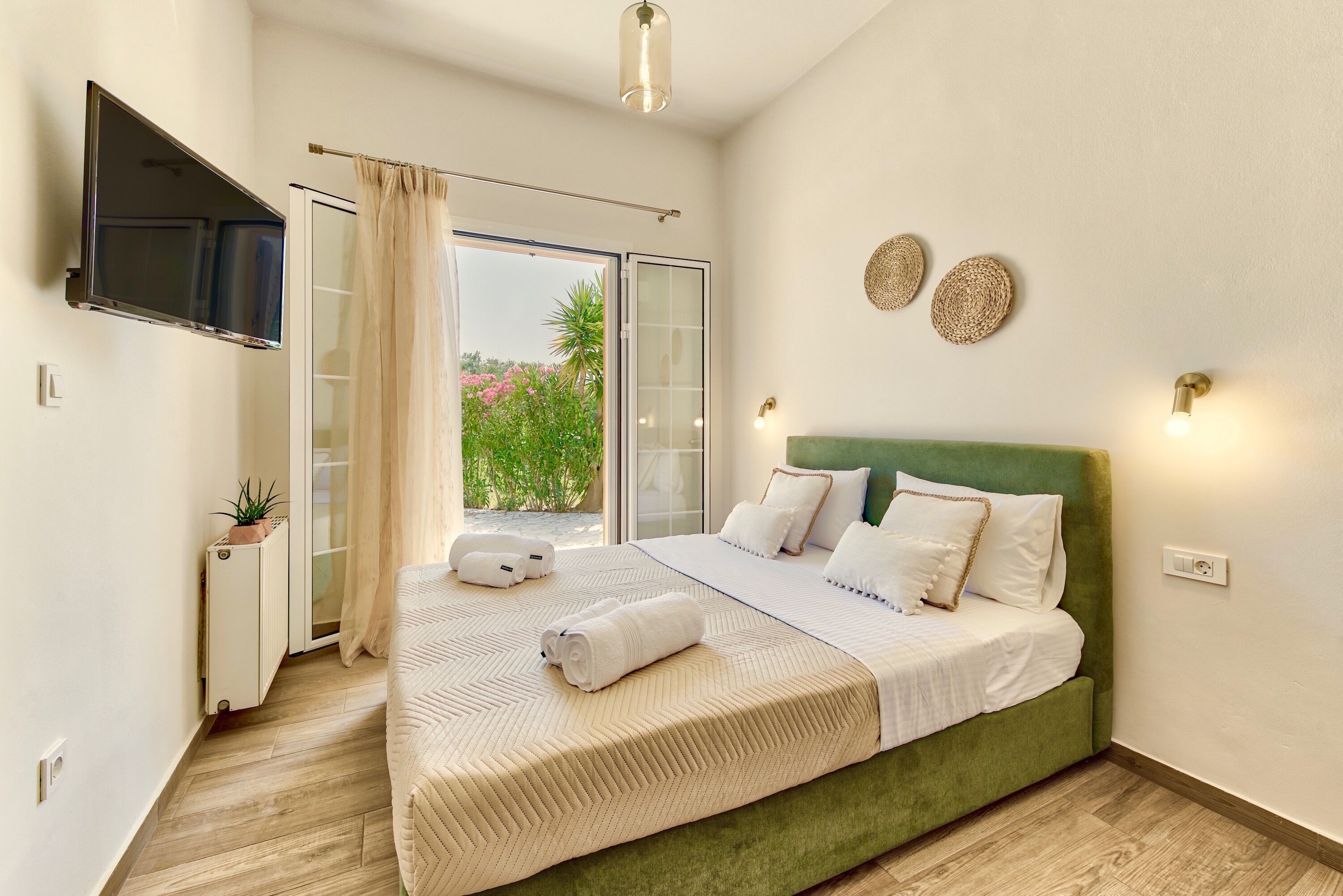 A bedroom with a green upholstered bed, a flat-screen TV mounted on the wall, a sliding glass door leading to a balcony, and a small bedside table with a plant.