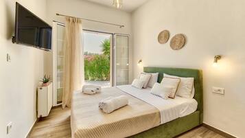 A bedroom with a green upholstered bed, a flat-screen TV mounted on the wall, a sliding glass door leading to a balcony, and a small bedside table with a plant.